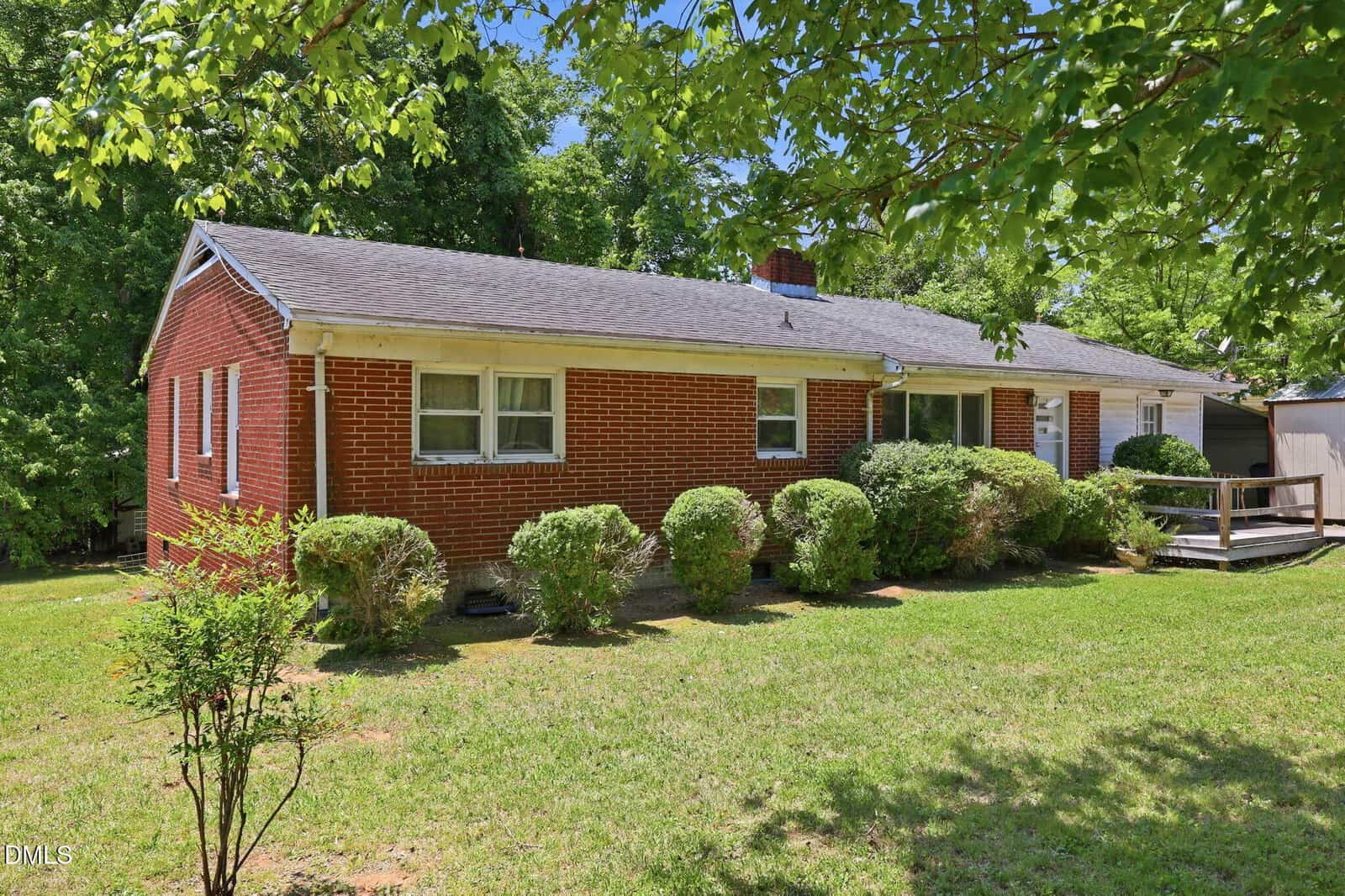A single-story red brick house with white trim and a gray shingled roof, surrounded by neatly trimmed bushes and trees, set on a grassy lawn with a ramp leading to the front entrance.