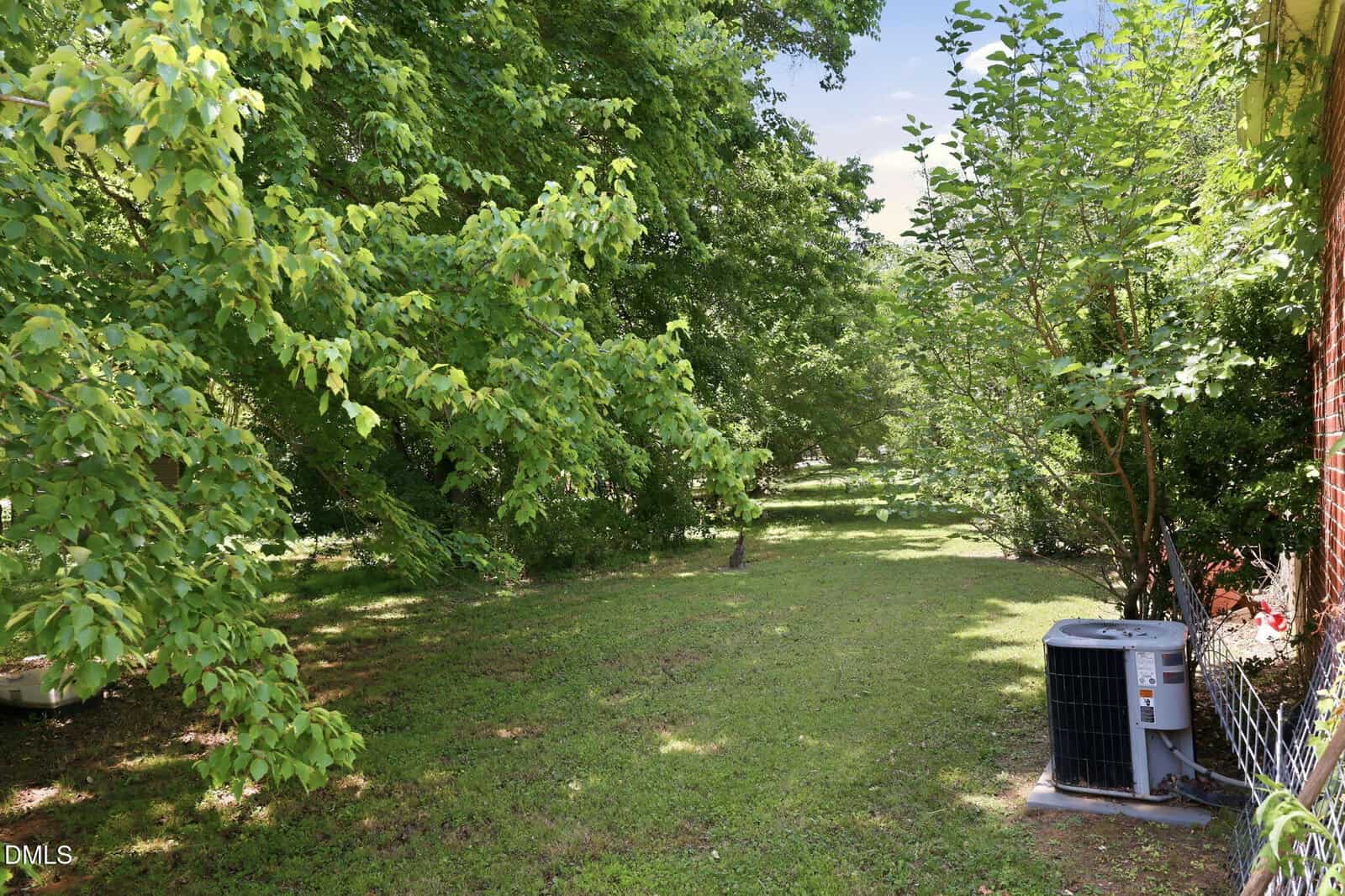 A grassy backyard with lush green trees providing shade, and an air conditioning unit is installed next to a brick house on the right. The area is bright and calm, with sunlight filtering through the trees.