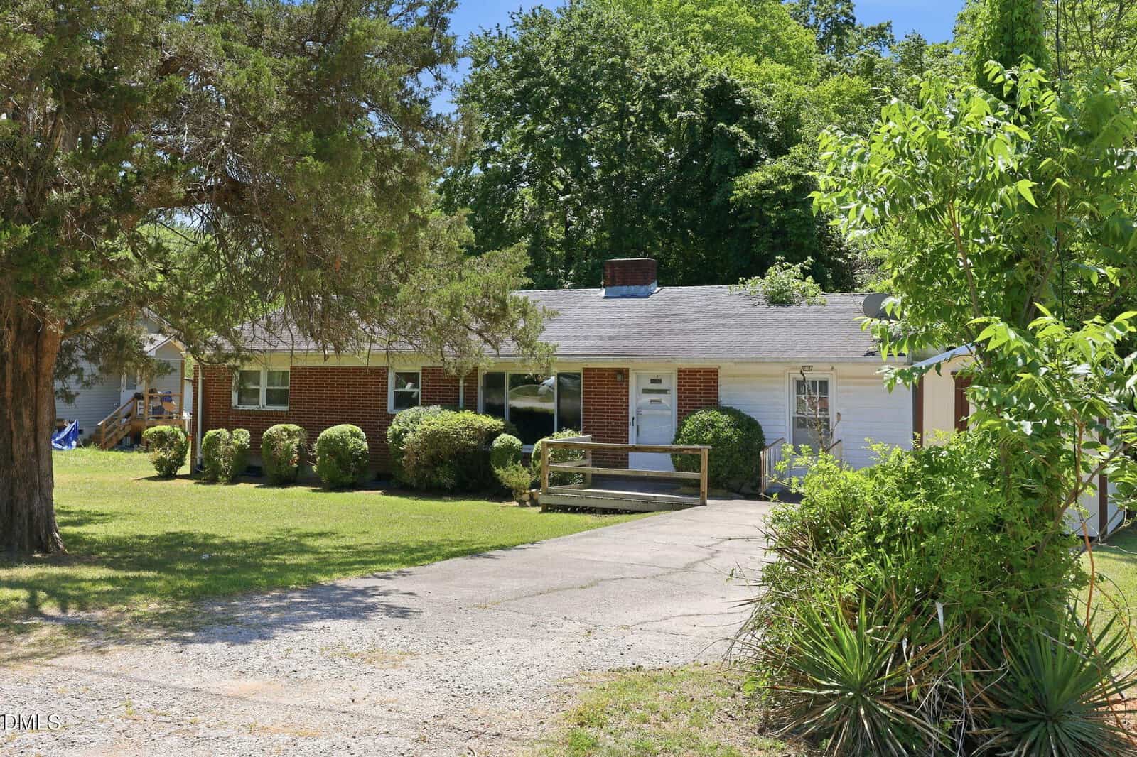 Single-story brick and white-sided house with a gray shingle roof, bushes along the front, a small front porch with a ramp, a grass lawn, and trees surrounding the property on a sunny day.
