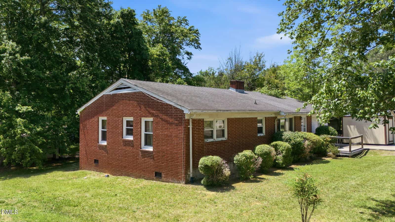 Single-story red brick house with multiple windows, a gray shingled roof, green shrubs along the side, and surrounded by grass and trees under a bright blue sky.