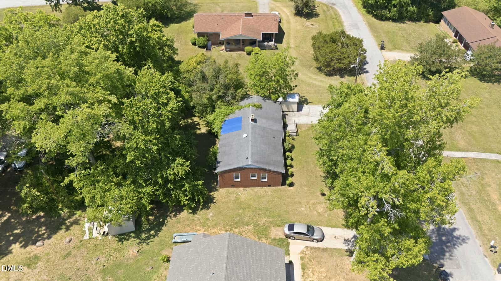 Aerial view of a brick house with a gray roof and blue tarp, surrounded by trees and neighboring homes, with a driveway and parked car in front.