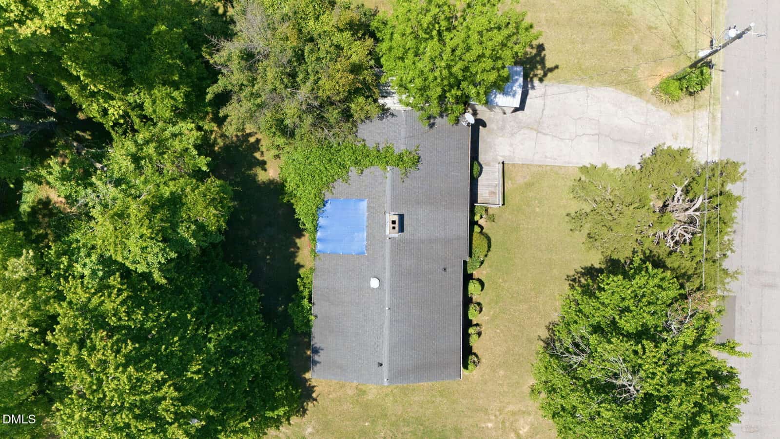 Aerial view of a single-story house with a gray roof, surrounded by green trees and a grassy yard. A driveway leads from the street to the house, and a small white structure is visible near the driveway.
