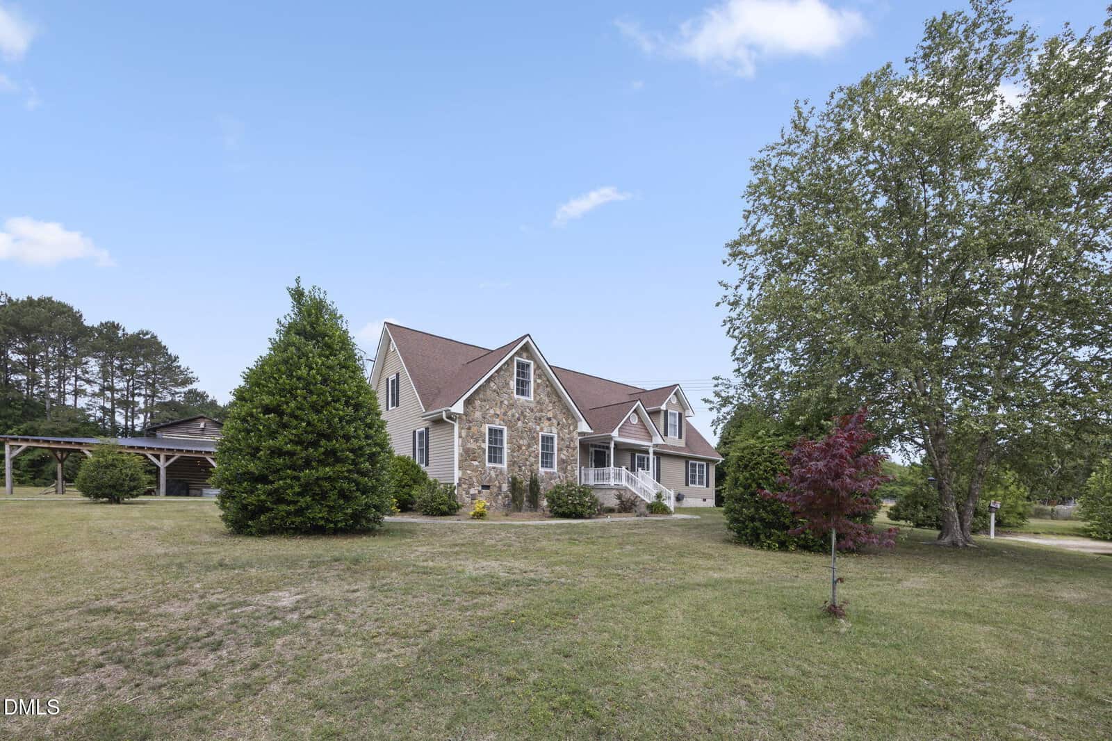 A two-story house with stone and beige siding, a brown roof, and multiple gables, sits on a spacious green lawn with several trees and a clear blue sky in the background.