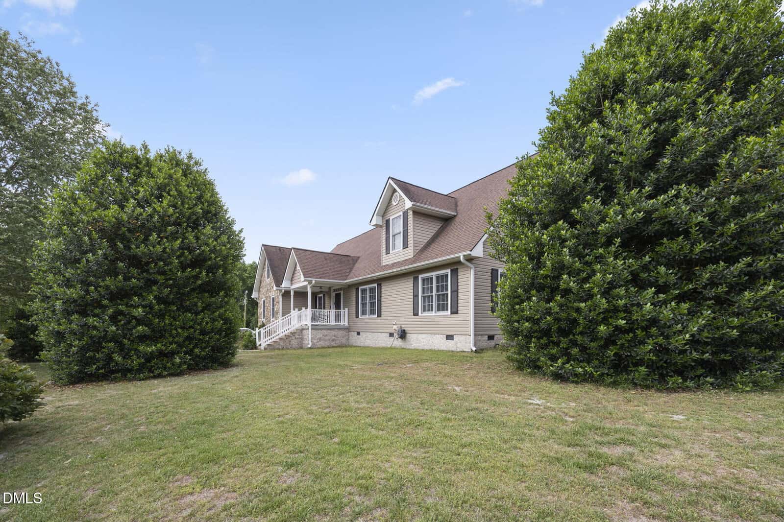 A beige house with a brown roof and white trim, surrounded by large green bushes and a grassy lawn under a blue sky.