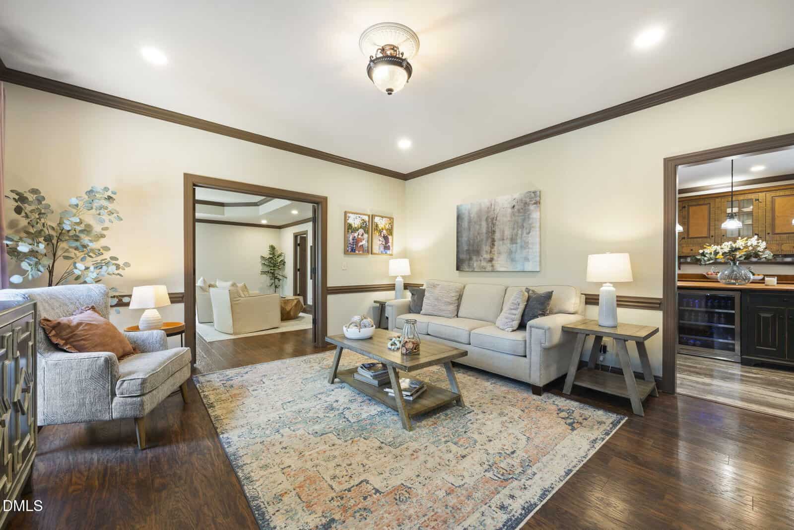 Bright living room with beige walls, a light sofa, armchair, coffee table, and patterned rug. Two lamps and framed artwork decorate the space. An open doorway leads to another seating area and a view of the kitchen.