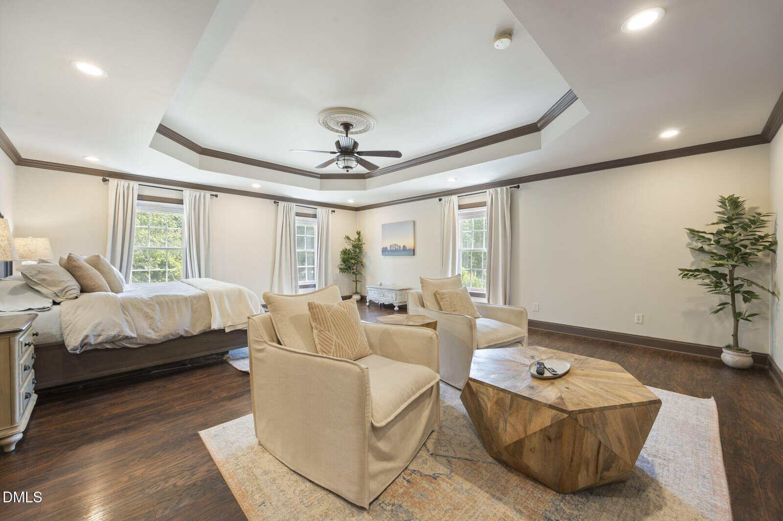 Spacious bedroom with a tray ceiling, ceiling fan, bed by three large windows with light curtains, two beige armchairs, a geometric wooden coffee table, and a potted plant on dark wood flooring.