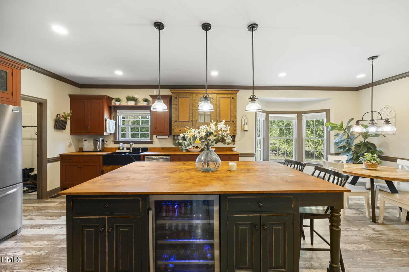 Spacious kitchen with a large wooden island, pendant lights, dark cabinets, stainless steel appliances, a dining area with a farmhouse table, and large windows letting in natural light.