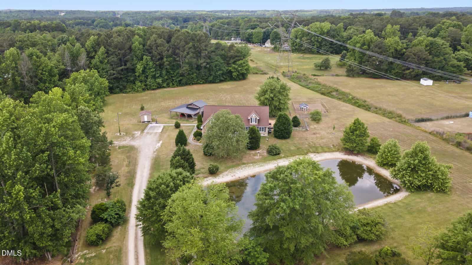 Aerial view of a large house with a brown roof, surrounded by trees and open fields, a gravel driveway, a small pond, and utility lines overhead in a rural setting.