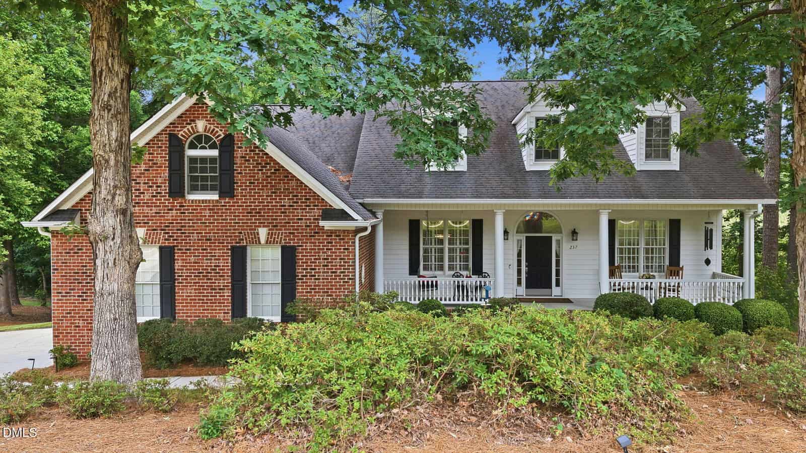 A two-story house at 237 Saddletree Road, Oxford, featuring a brick and white exterior, spacious front porch with columns, dormer windows, and a gabled roof, all surrounded by trees and landscaped shrubs.