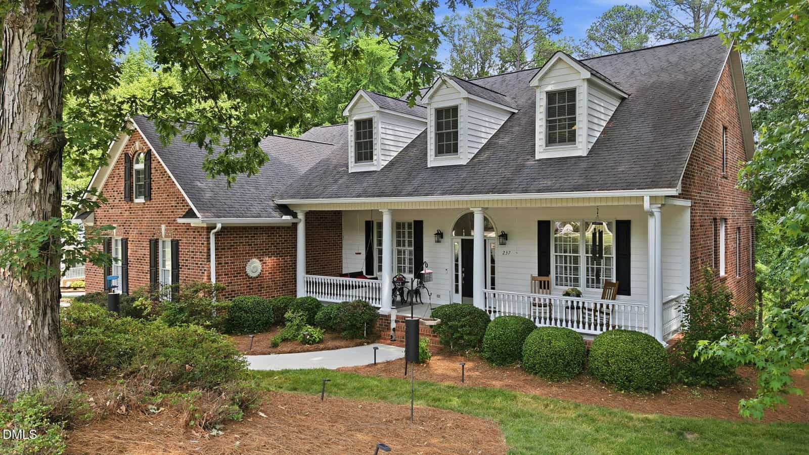 A brick and white-paneled house with dormer windows, a large covered front porch, white railings, and a manicured yard with shrubs and trees. The house is surrounded by lush greenery.