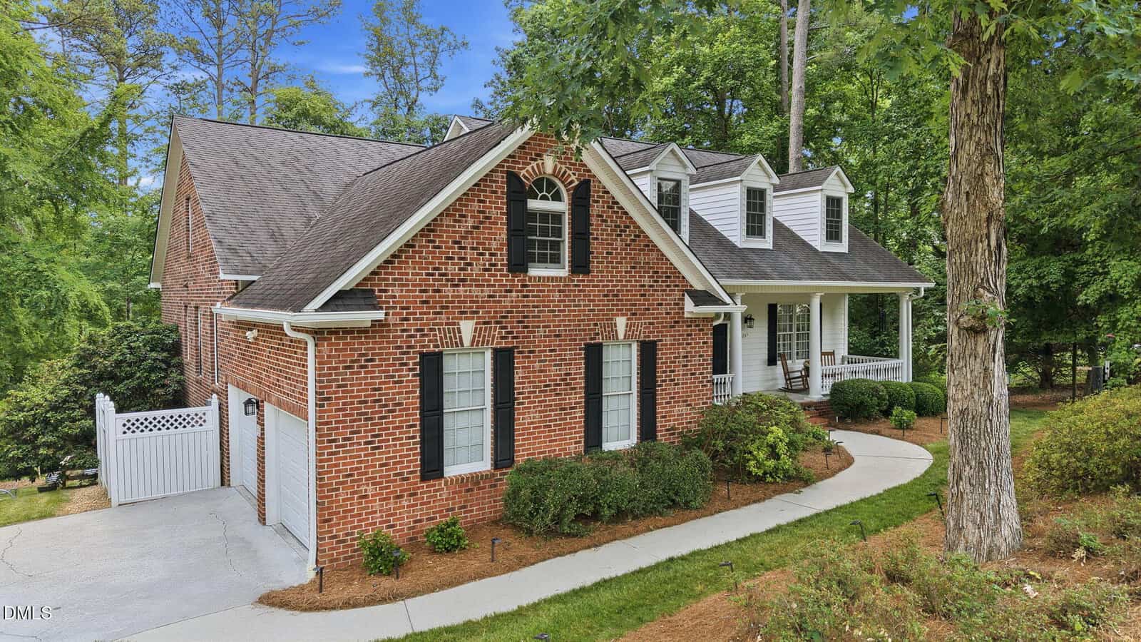 A red-brick house with white trim and black shutters, featuring a covered front porch with columns, dormer windows, and surrounded by green trees and landscaped shrubs. A curved driveway leads to the entrance.
