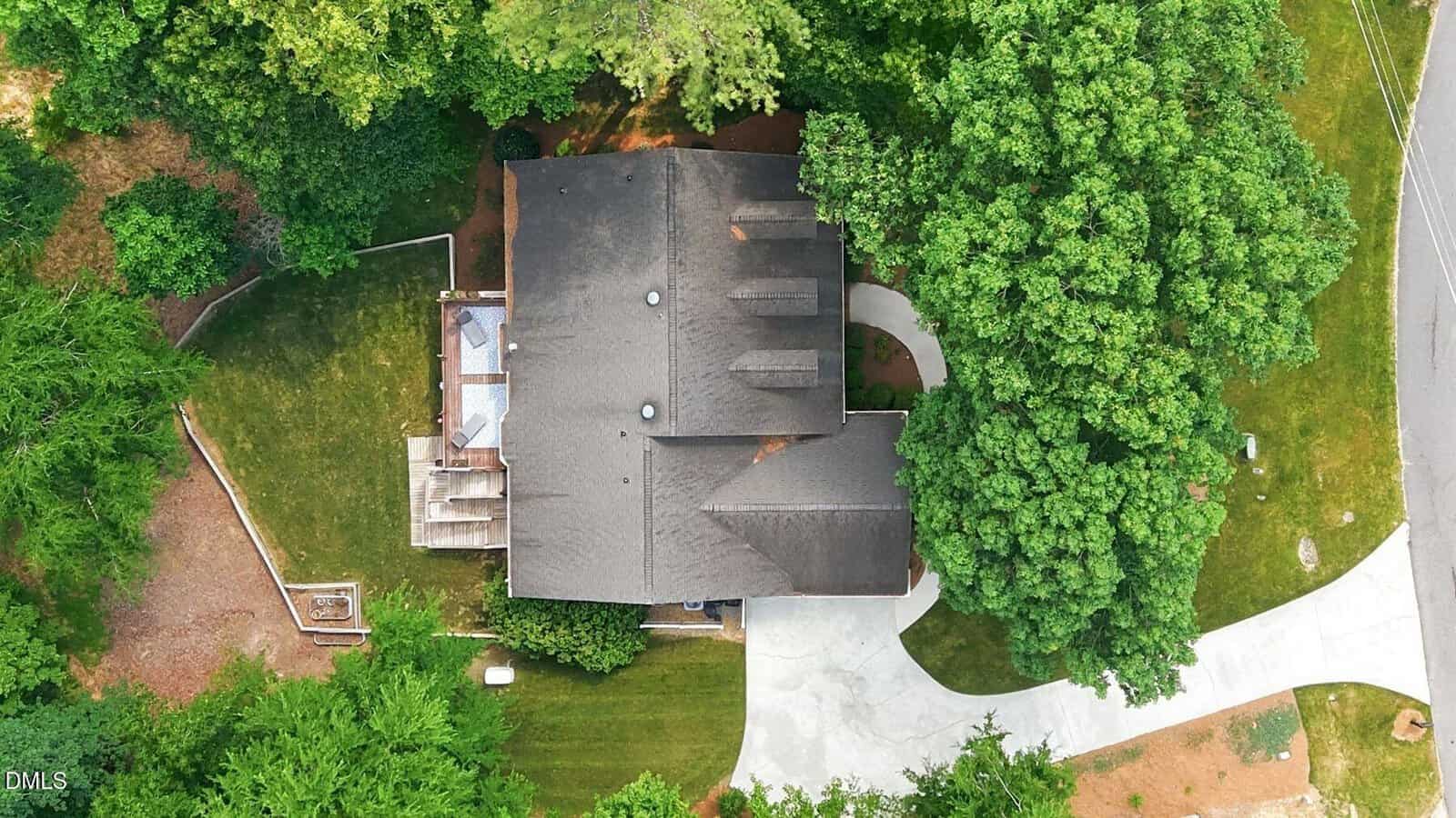 Aerial view of a house surrounded by trees with a curved driveway, deck area on the left, and well-maintained lawn. The roof is dark, and the property is bordered by a street on the right.