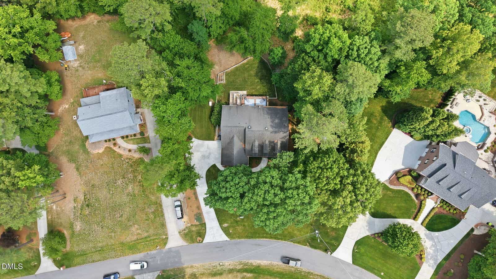Aerial view of a residential area featuring three houses surrounded by trees. One house is centered with a driveway and backyard, while the other homes and a swimming pool are nearby. Cars are parked along the street and in driveways.