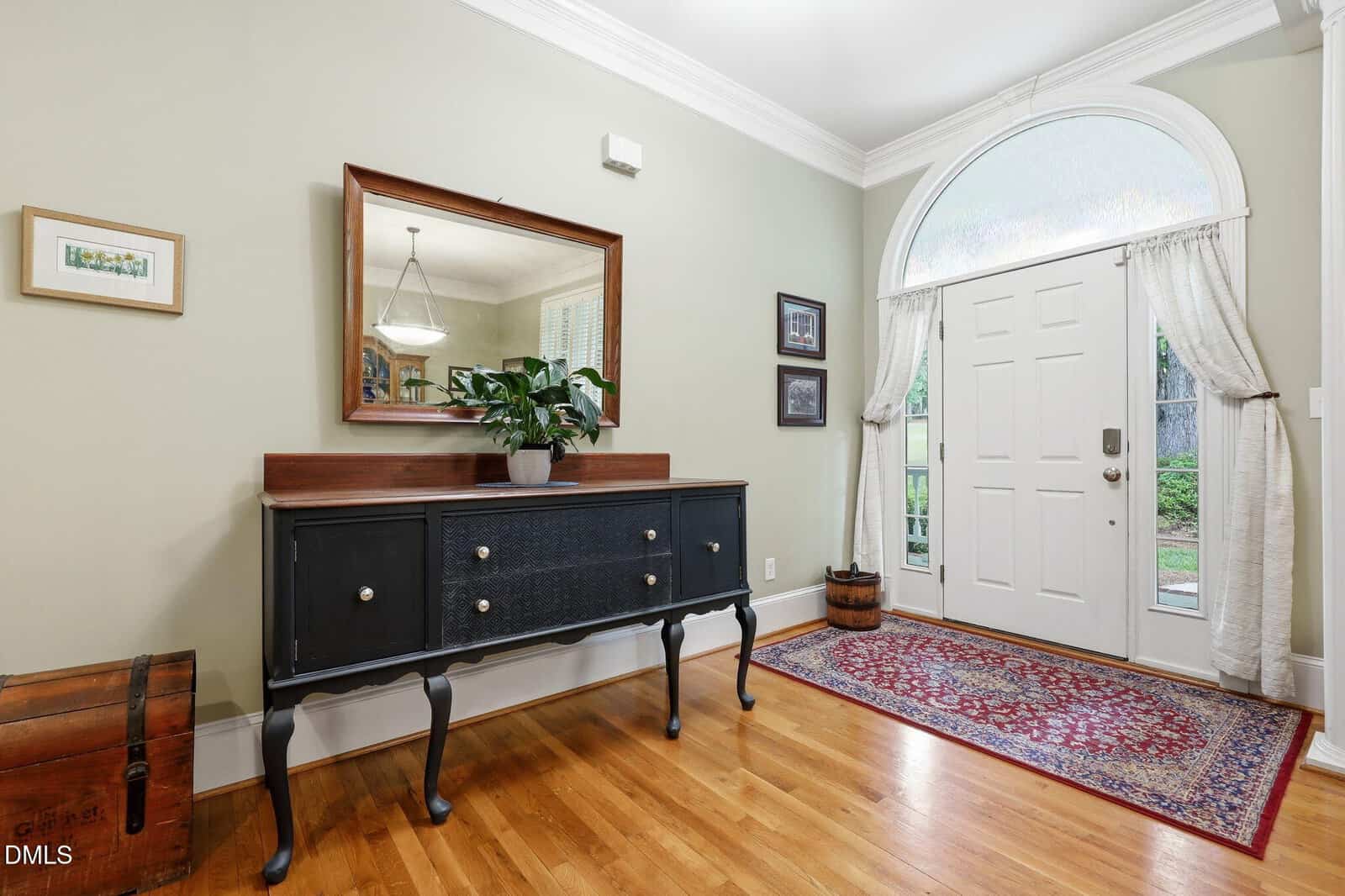 Bright entryway with hardwood floors, a black sideboard holding a potted plant, a large mirror above, framed art on the walls, a red rug, and a white front door with glass arch and sheer curtains.