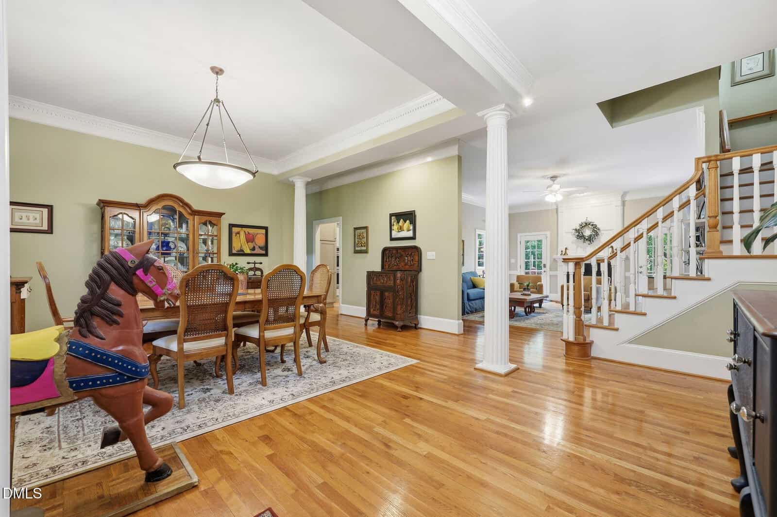 Spacious dining area with wooden floors, a carousel horse, and a staircase leading upstairs. A china cabinet, dining table, and chairs sit near green walls. Living room visible in the background.
