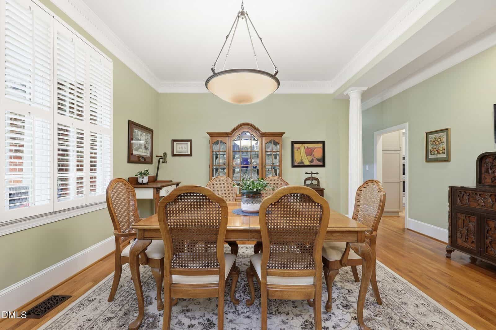 A dining room with a wooden table, six chairs, a chandelier, a hutch, and light green walls. A potted plant sits on the table, and framed art decorates the walls. Large windows with shutters let in natural light.