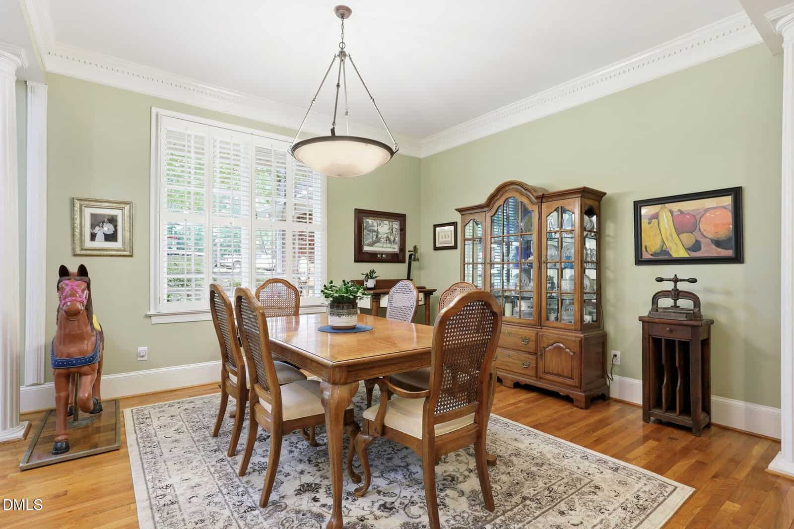 Traditional dining room with a wooden table, six chairs, china cabinet, area rug, window with blinds, wall art, and a decorative rocking horse in the corner on hardwood floors.