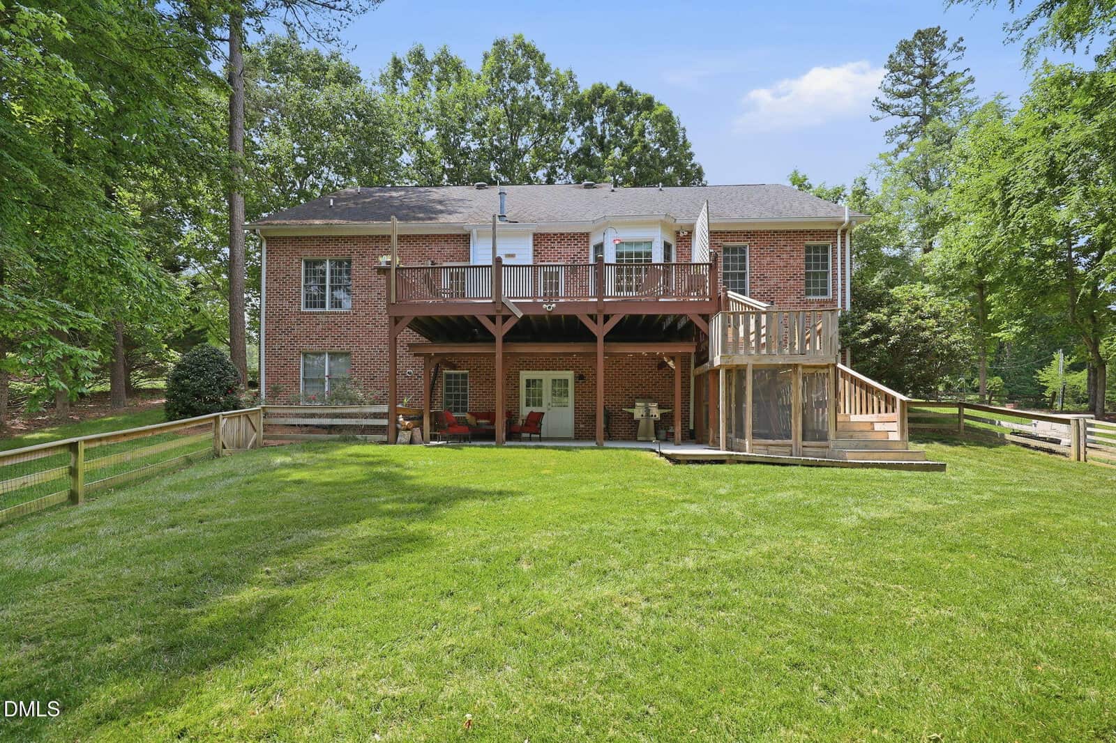 Two-story brick house with a large elevated wooden deck and covered patio area underneath, set in a spacious fenced backyard with green grass and surrounded by tall trees.