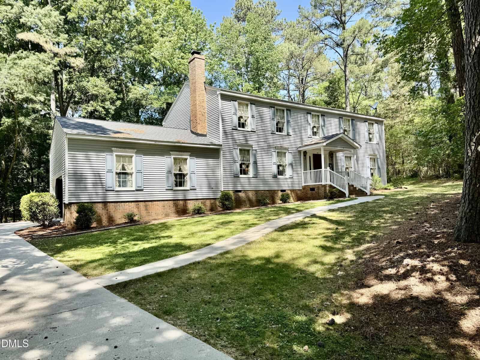 Located at 84 Wintergreen Road in Henderson, this two-story house features light gray siding, white trim, a brick foundation, and a covered front entrance. Multiple windows with shutters overlook a sloped lawn surrounded by trees.