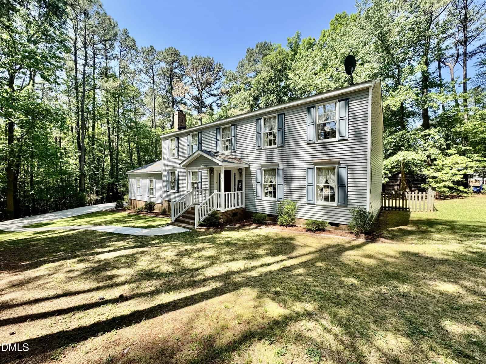 Two-story gray house with white trim, shutters, and a front porch, surrounded by trees and a spacious grassy yard on a sunny day.