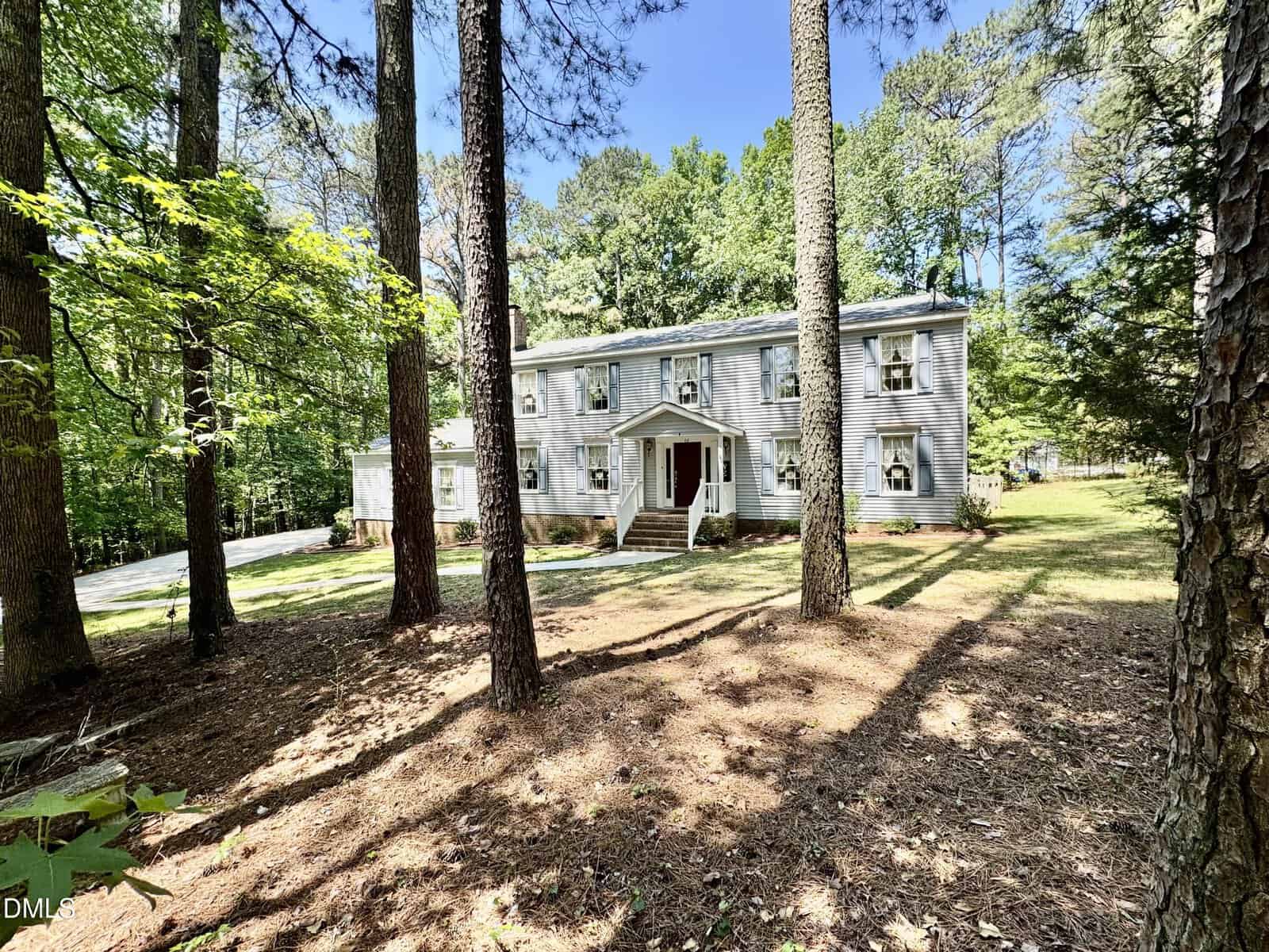 A two-story white house with shuttered windows and a small front porch sits among tall trees and a pine-covered yard on a sunny day. The driveway curves along the left side of the property.