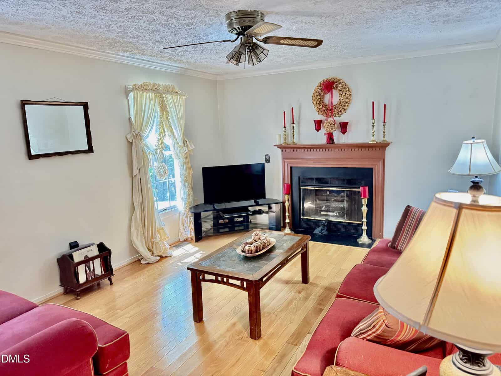 Bright living room with wood floor, red sofas, wooden coffee table, fireplace decorated with candles and a wreath, TV in the corner, standing lamp, window with drapes, and ceiling fan overhead.