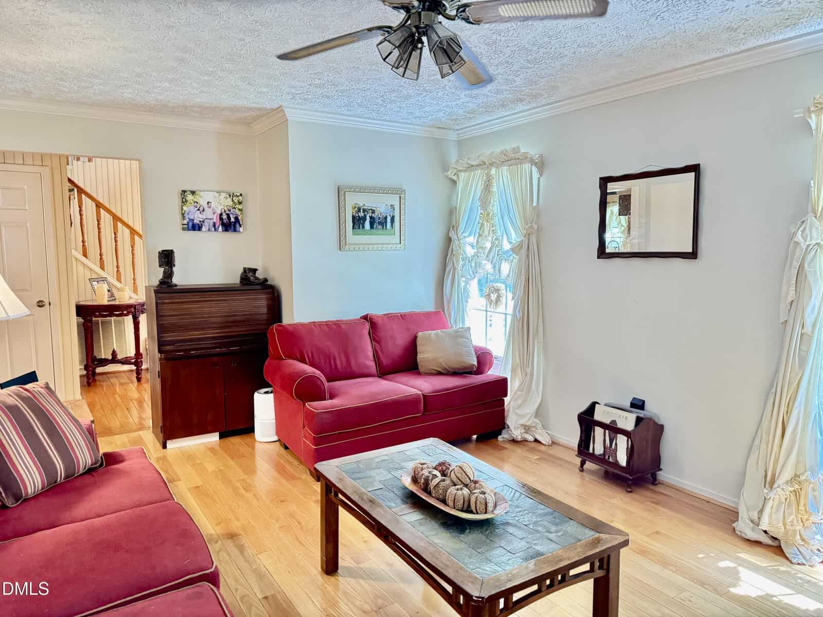 Bright living room with red sofas, a wooden coffee table, ceiling fan, piano, framed art, and large windows with white drapes letting in natural light. Hardwood floors and light walls give the space an airy feel.