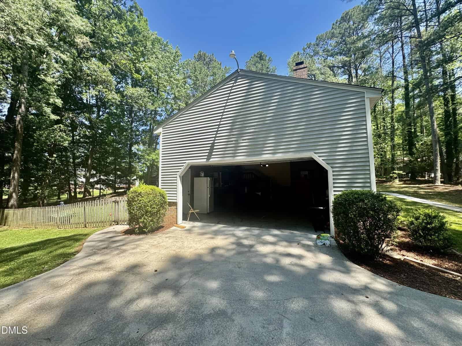 A two-car garage with gray siding is open, revealing a spacious interior. The driveway is surrounded by green shrubs and tall trees, with a fenced yard visible to the left under a clear blue sky.