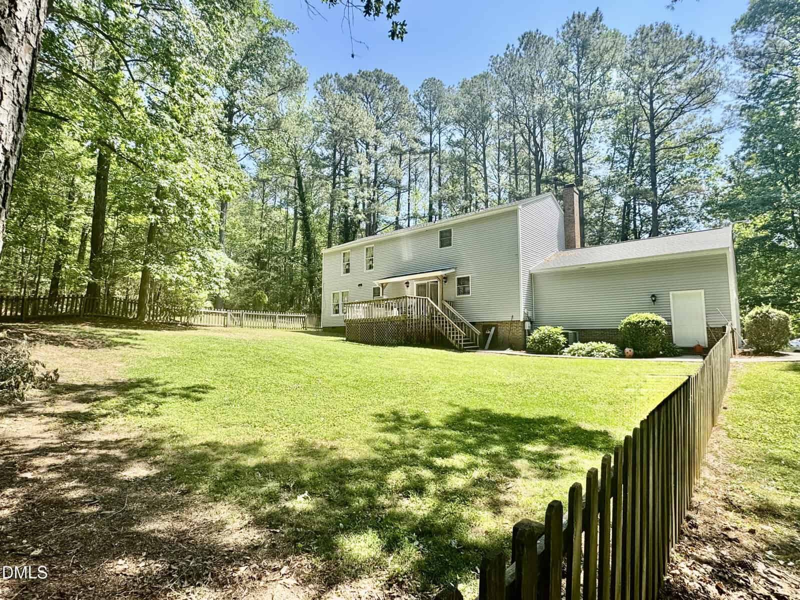 A two-story light gray house with a wooden deck and fenced backyard, surrounded by green grass, trees, and dense forest under a bright blue sky.