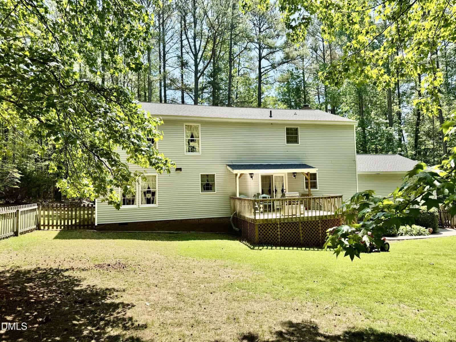A two-story white house with a wooden deck in a grassy backyard, surrounded by trees and partial fencing, on a sunny day.
