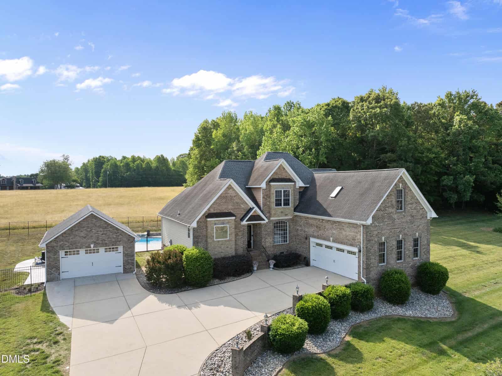 Aerial view of a large brick house at 120 Barn Hill Lane, Wake Forest, with multiple gables and a three-car garage, set on a spacious lot with manicured landscaping, detached garage, backyard pool, and scenic views of trees and open fields.