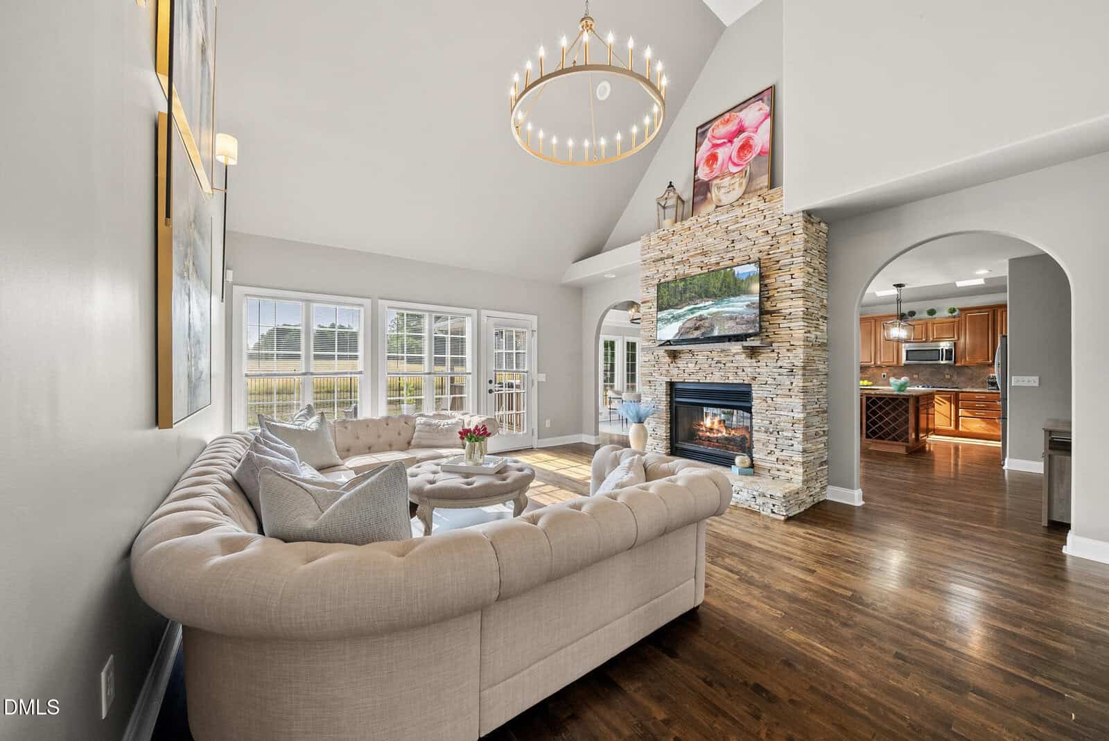 A spacious living room with a large beige sectional sofa, a round ottoman, a stone fireplace with a TV above it, wooden floors, high ceilings, and tall windows letting in natural light. The kitchen is visible through an archway.