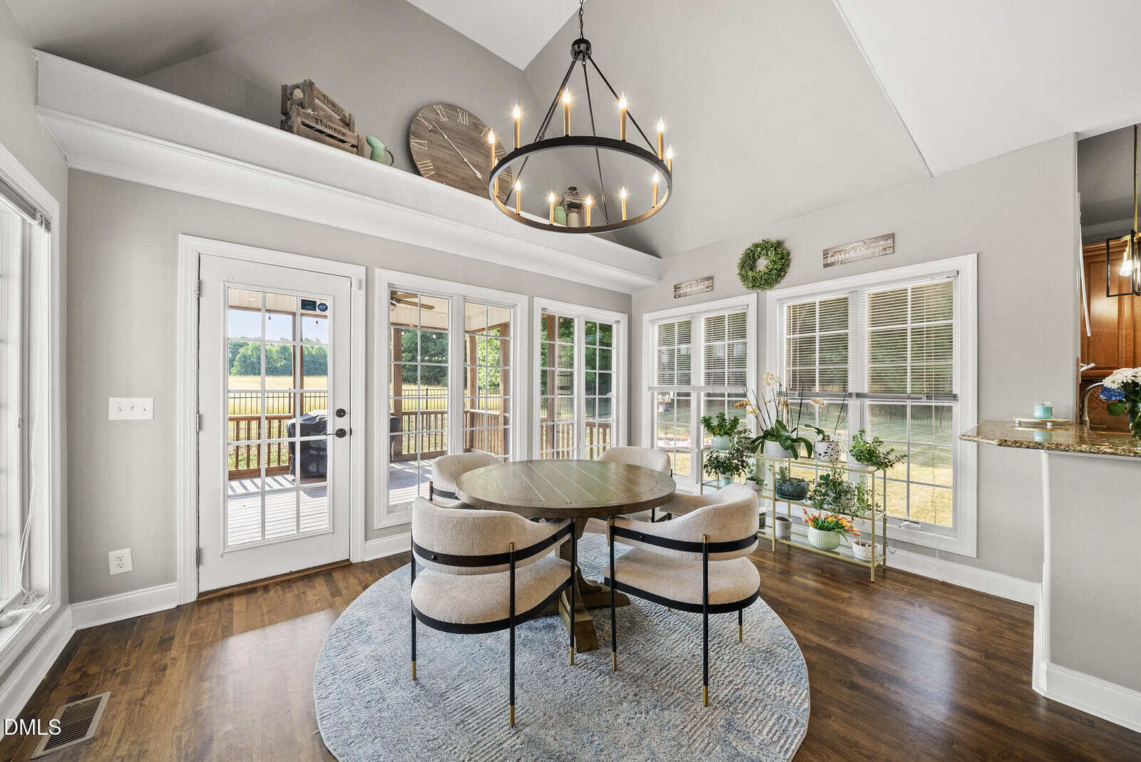 Bright dining area with round table and four chairs on blue rug, large windows and glass door showing outdoor view, chandelier overhead, and decorative plants and wall accents throughout the room.