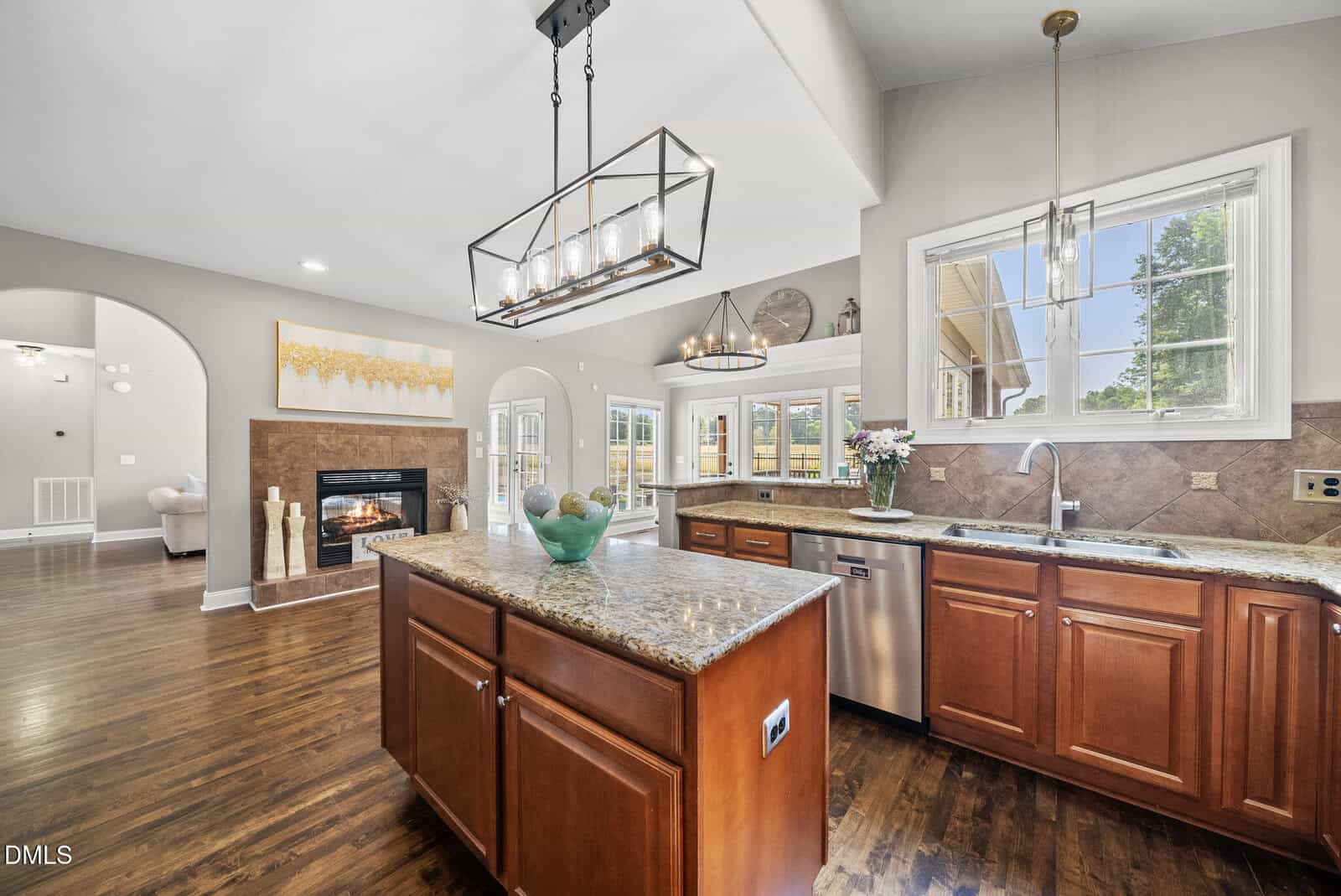 Spacious kitchen with granite countertops, wood cabinets, a center island, stainless steel appliances, large windows, and pendant lighting. The kitchen opens to a living area with a fireplace and hardwood floors.