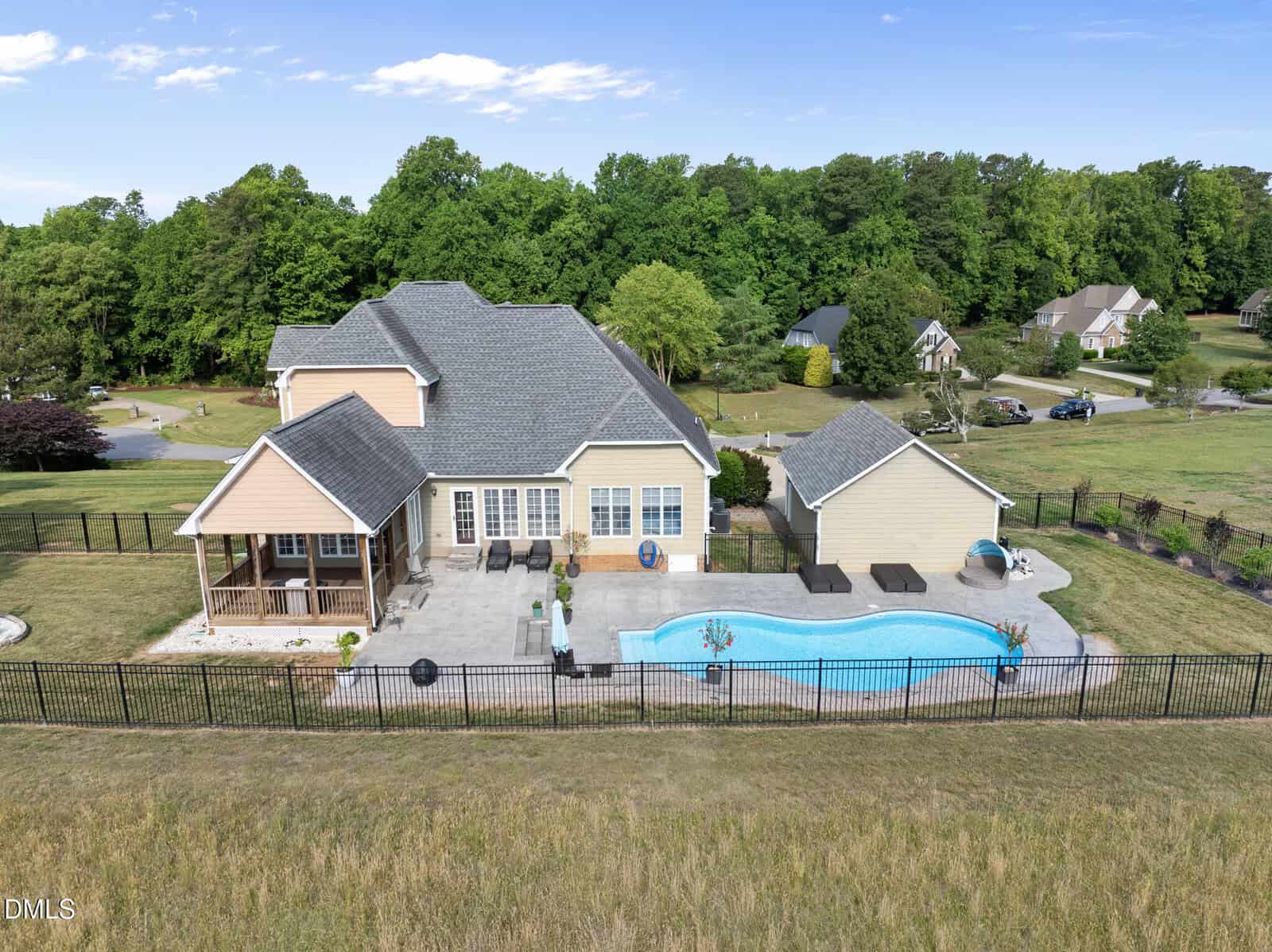 A large suburban house with a fenced backyard, patio seating, and an in-ground swimming pool, surrounded by green lawns and trees, with neighboring homes in the background.