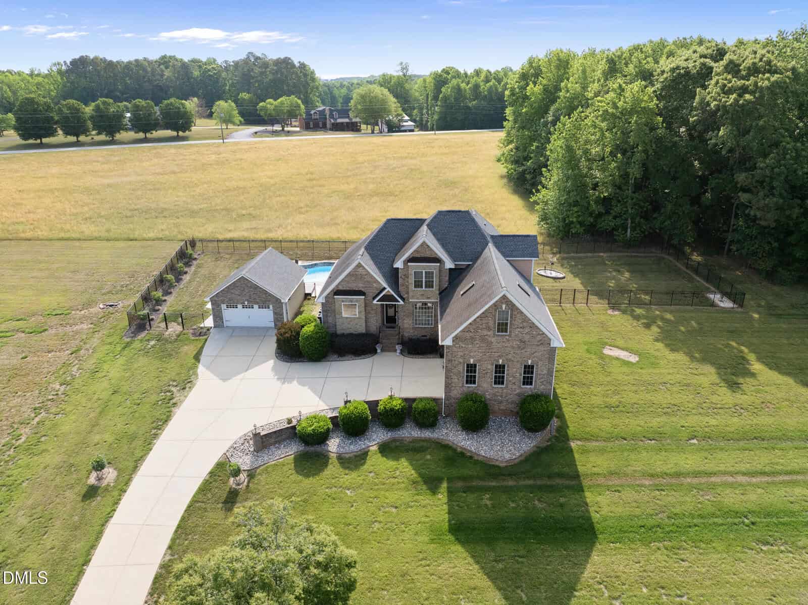 Aerial view of a large brick house with a detached garage, pool, and long driveway, surrounded by fields, trees, and a fenced backyard on a sunny day.