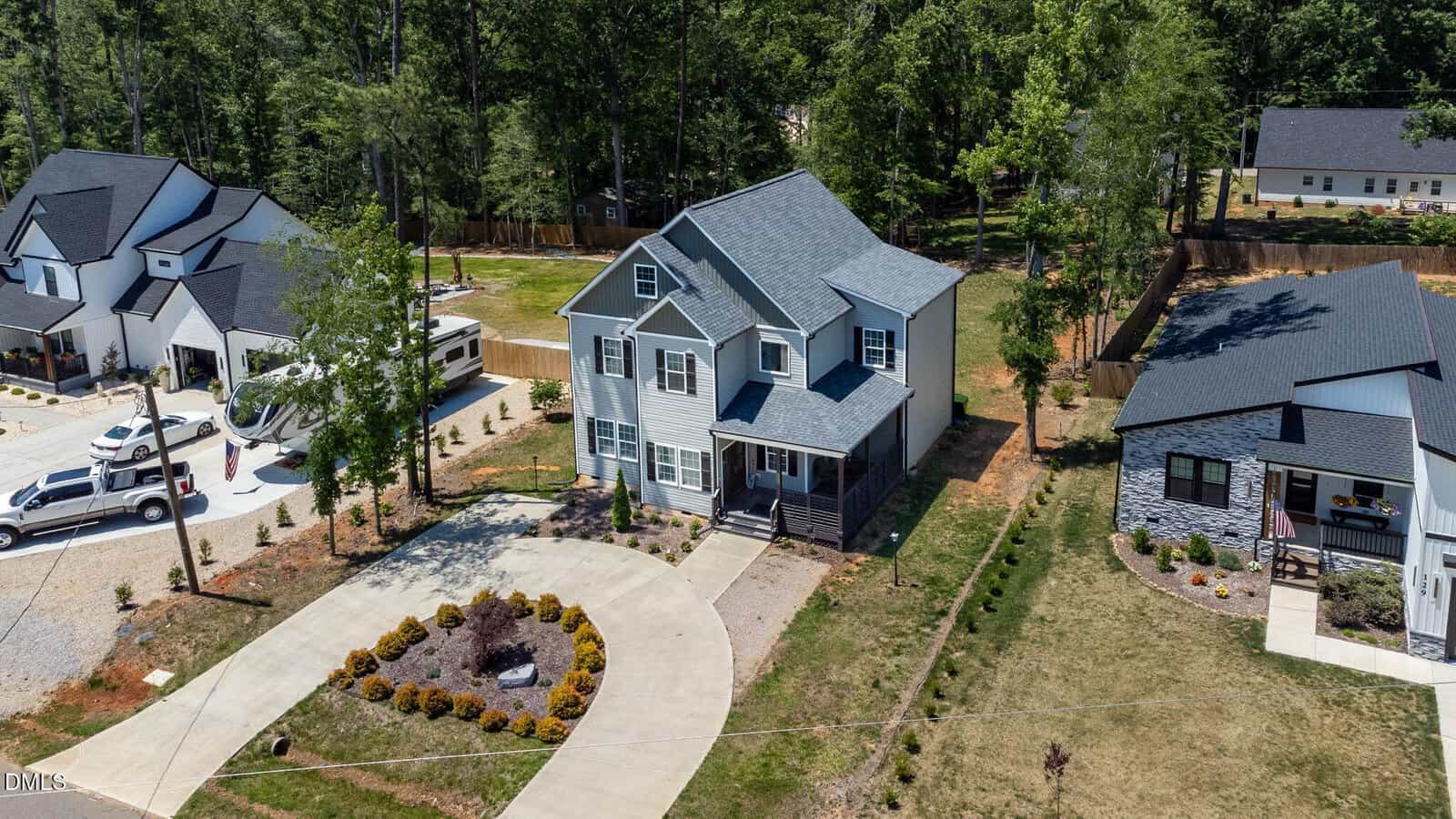Aerial view of a two-story gray house with a covered front porch, circular driveway, landscaped front yard, and neighboring houses surrounded by trees.