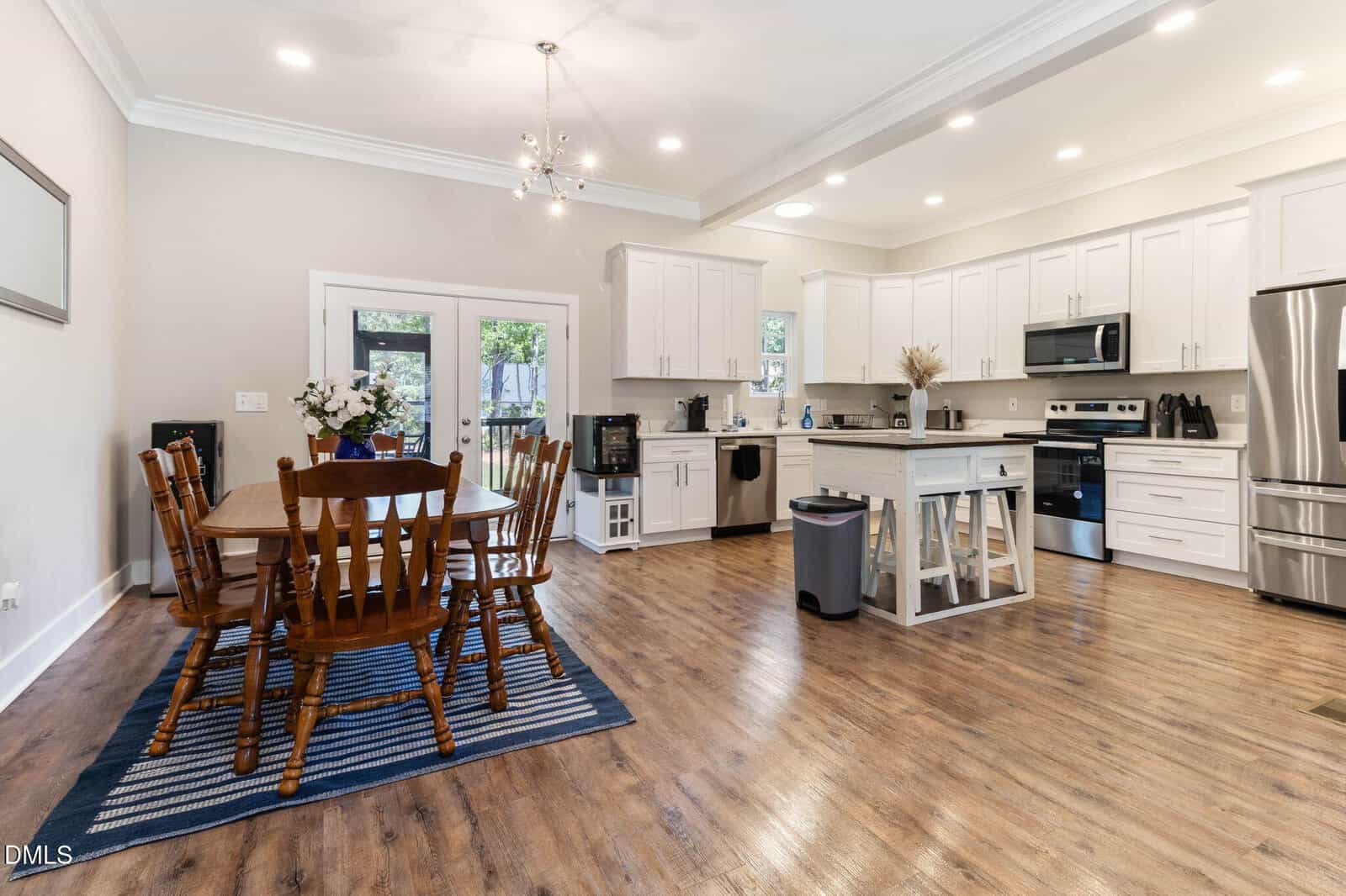 Spacious kitchen with white cabinets, stainless steel appliances, and a center island. Wooden dining table with six chairs sits on a blue rug. Hardwood floors and recessed lighting brighten the room.