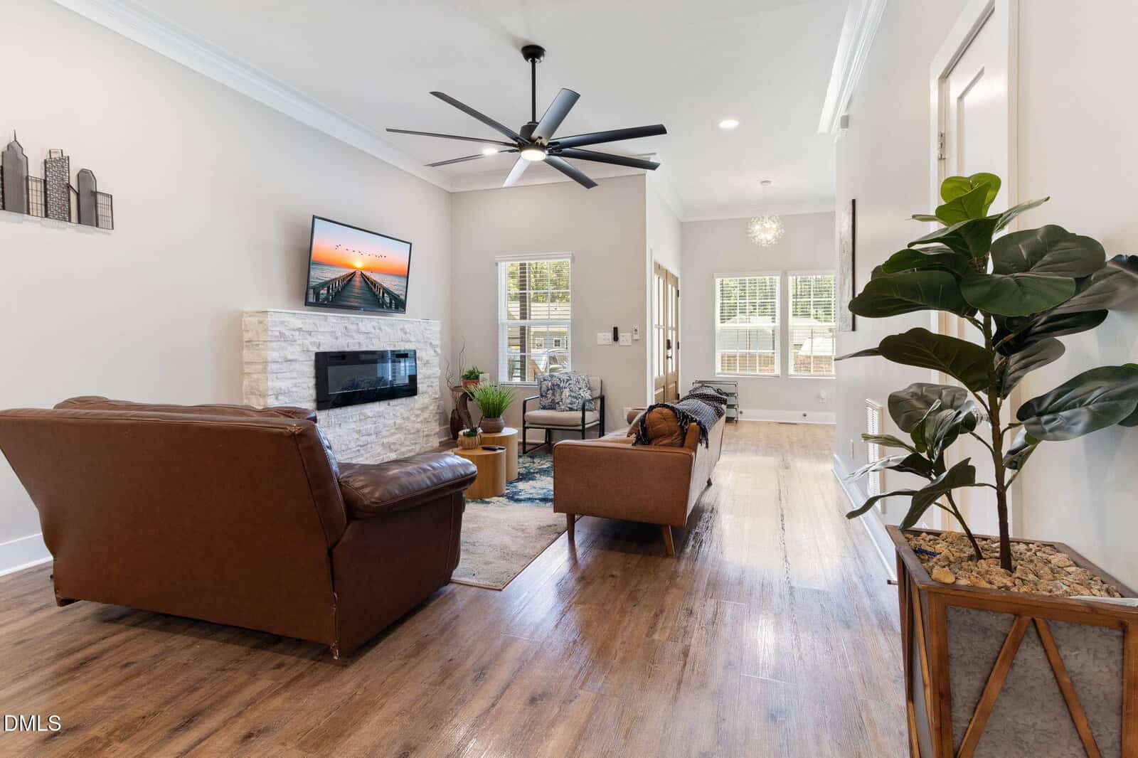Modern living room with wood flooring, brown leather sofas, a potted plant, wall-mounted TV above a white stone fireplace, and large windows letting in natural light.