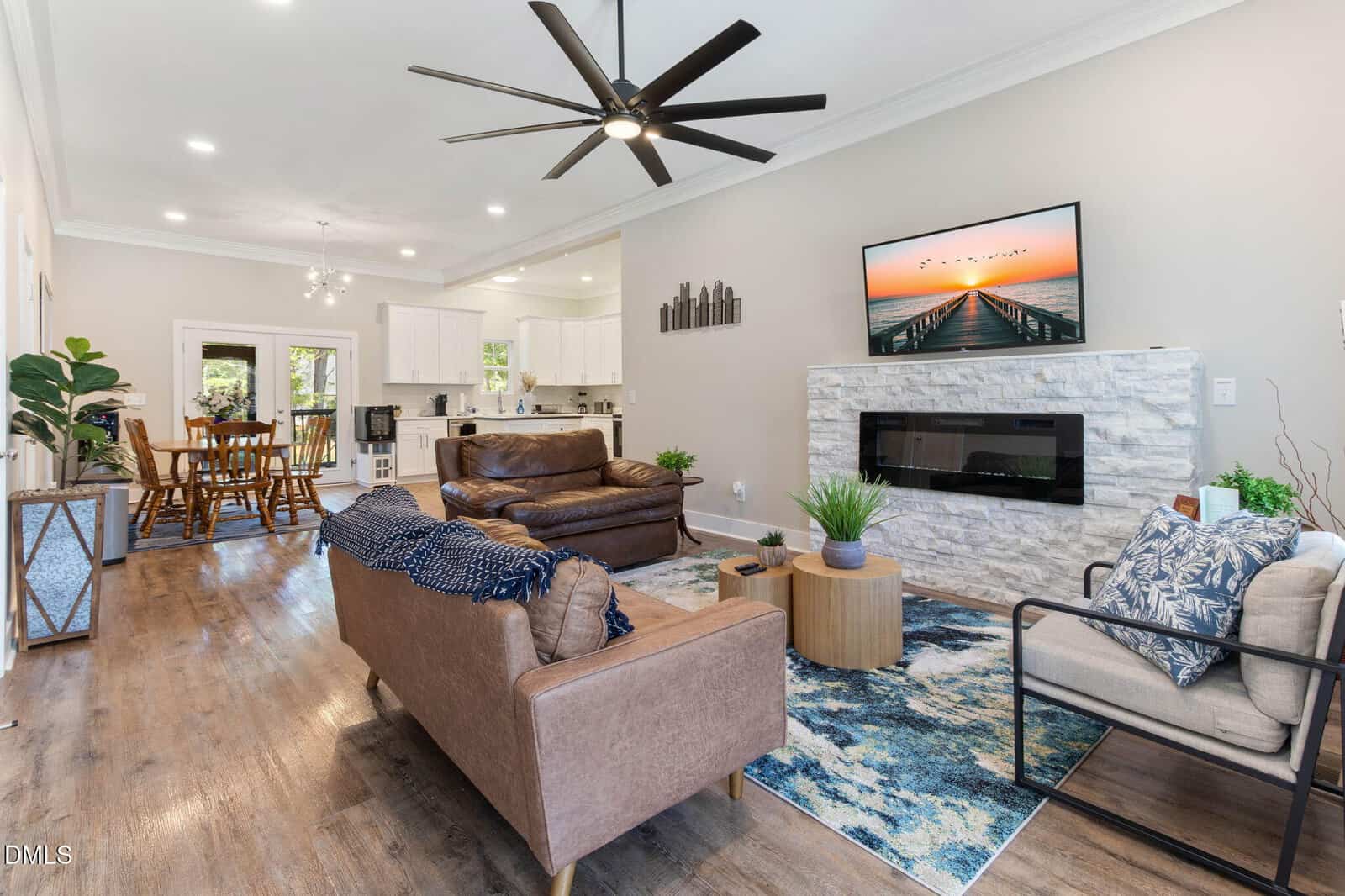 Modern living room at 127 Ottawa Drive in Louisburg with a brown leather sofa, gray couch, armchair, blue rug, coffee table, stone fireplace, lush plants, and large ceiling fan. Open layout connects to dining area and white kitchen.