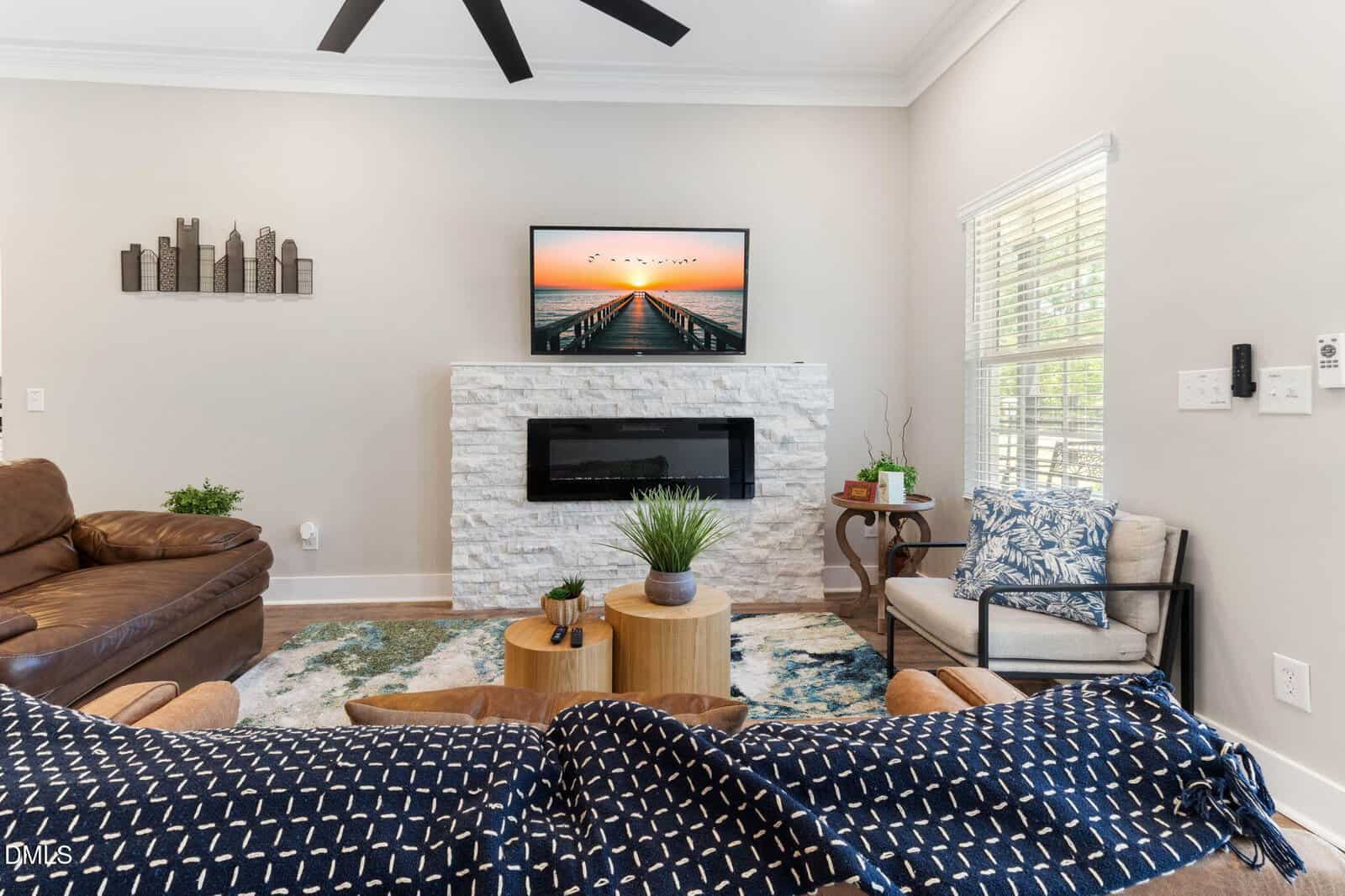 Modern living room with a stone fireplace, wall art, TV above the mantel, brown leather sofa, armchair, plants, and a large window letting in natural light. Blue patterned throw covers the foreground.