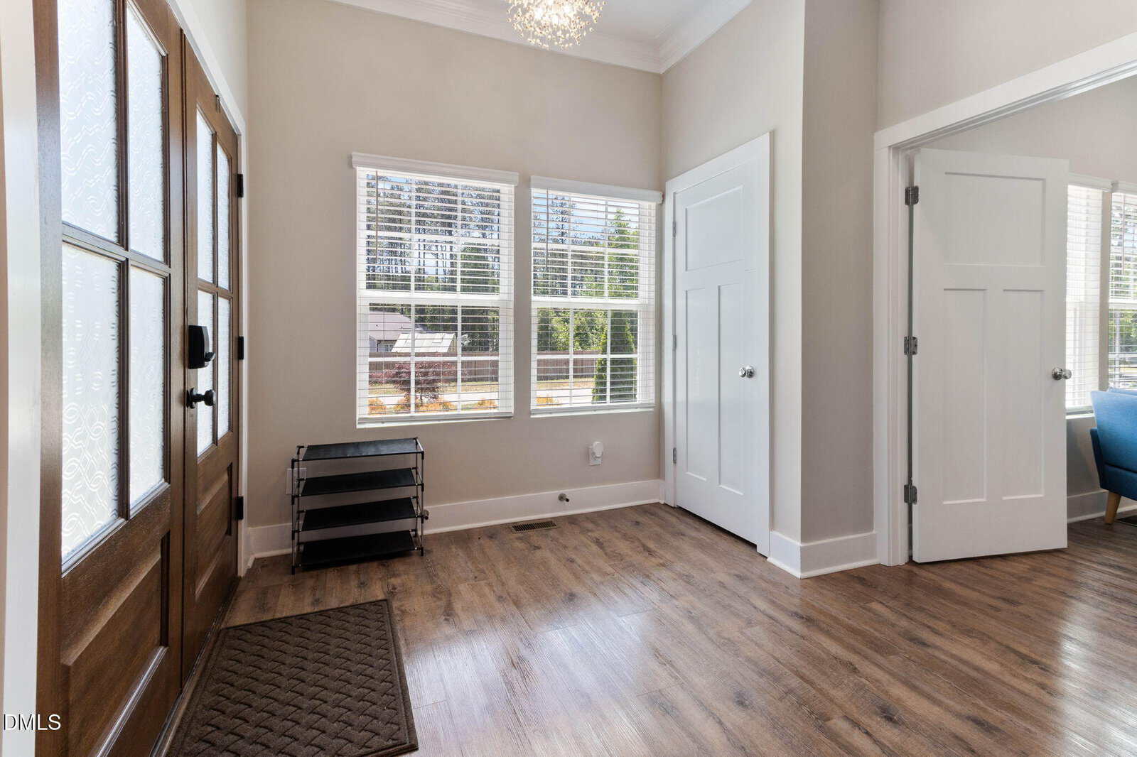 Bright entryway with wood floors, double wooden front doors with glass panels, a black mat, metal shoe rack, and two large windows letting in natural light. A doorway leads to a room with blue furniture.