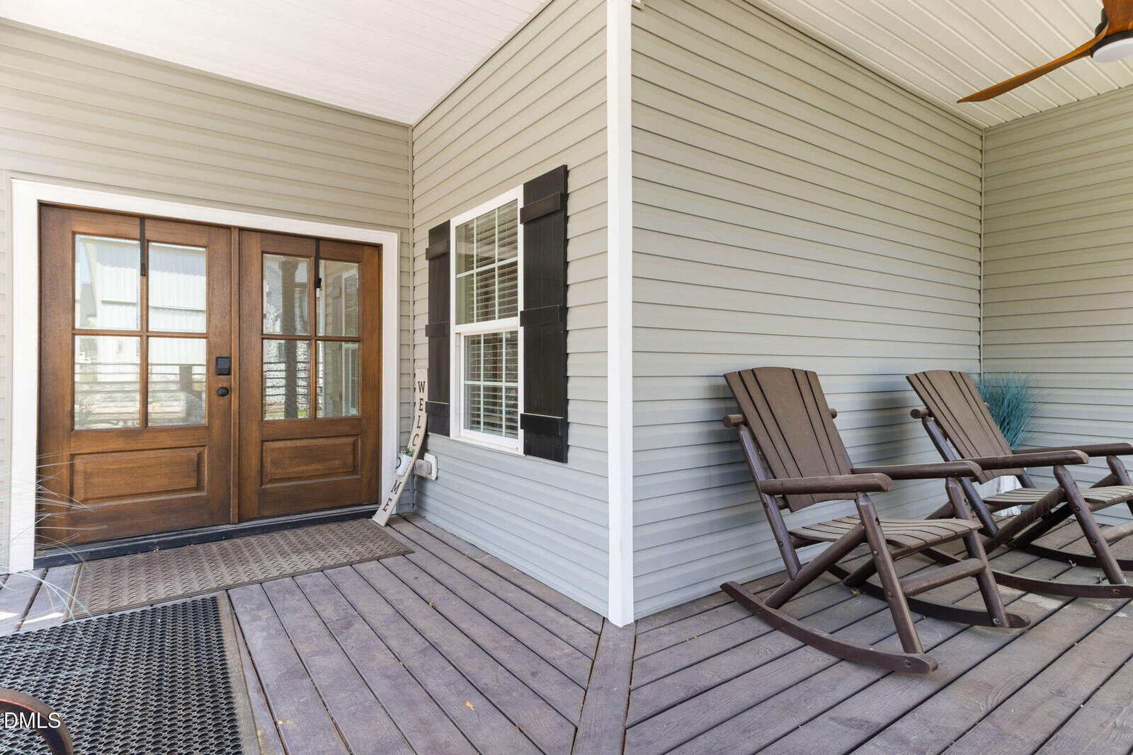 A porch with wooden flooring features two brown rocking chairs, a small table, a welcome sign, a double wooden front door with glass panes, and light gray siding on the house.