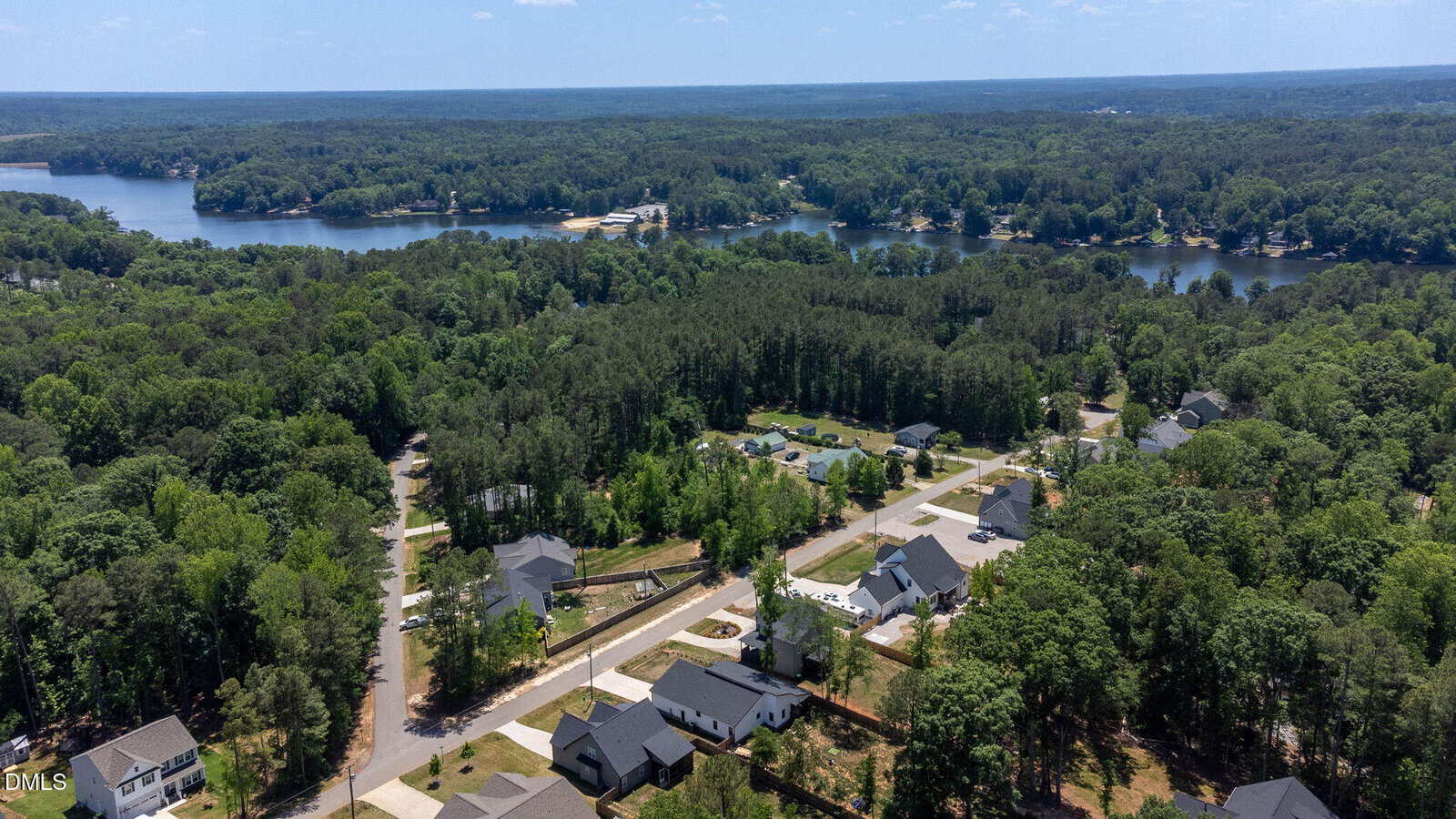 Aerial view of a suburban neighborhood surrounded by dense green forest, with a lake and tree-lined shoreline in the background under a clear blue sky.