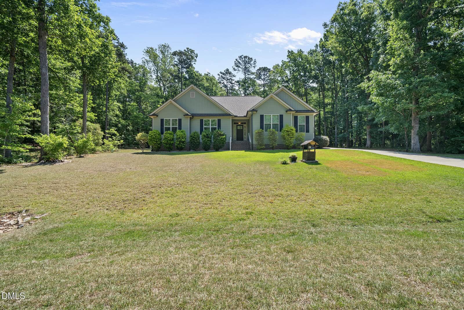 A single-story green house with white trim sits in the center of a large, grassy yard surrounded by tall trees under a clear blue sky. A driveway is visible on the right side.