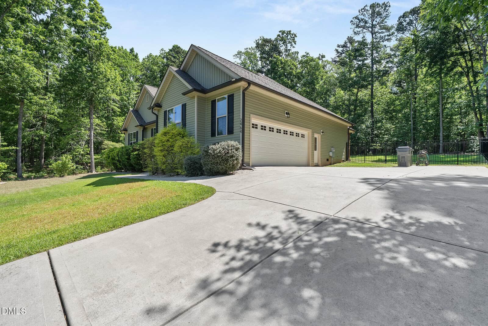 Single-story house with light green siding, gable roof, and attached two-car garage. A wide concrete driveway leads up to the home, surrounded by green lawn, shrubs, and trees under a clear sky.