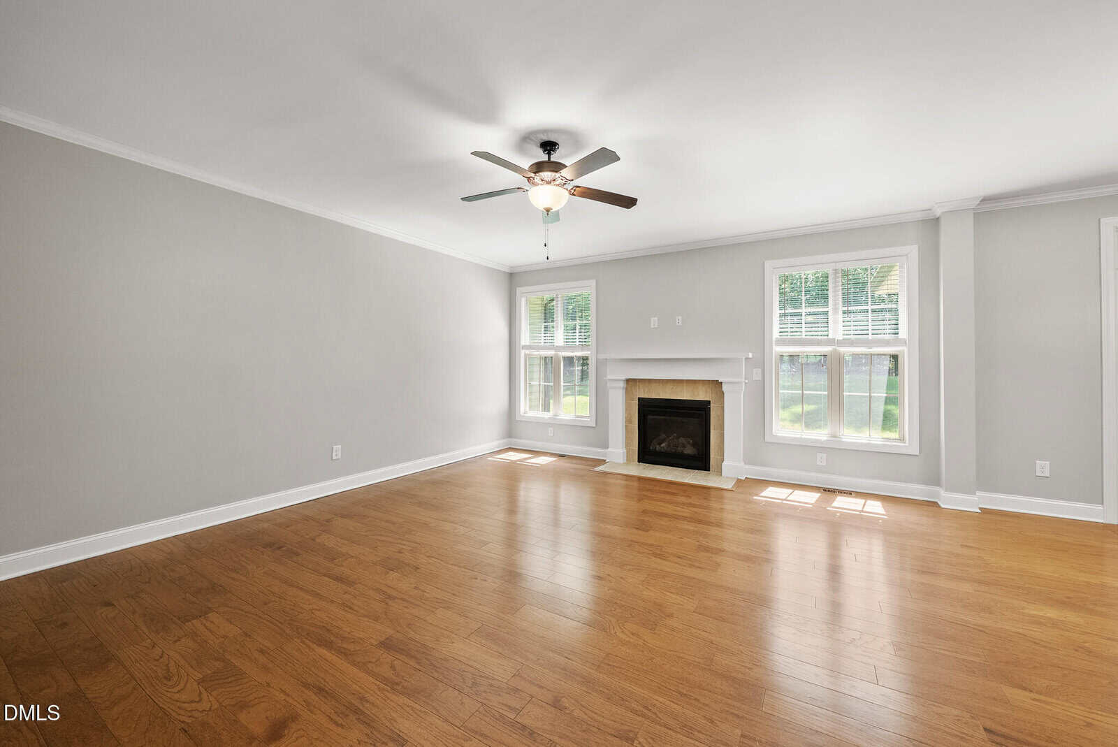 Spacious, empty living room with light gray walls, wood flooring, a ceiling fan with light, a white fireplace, and large windows letting in natural light.