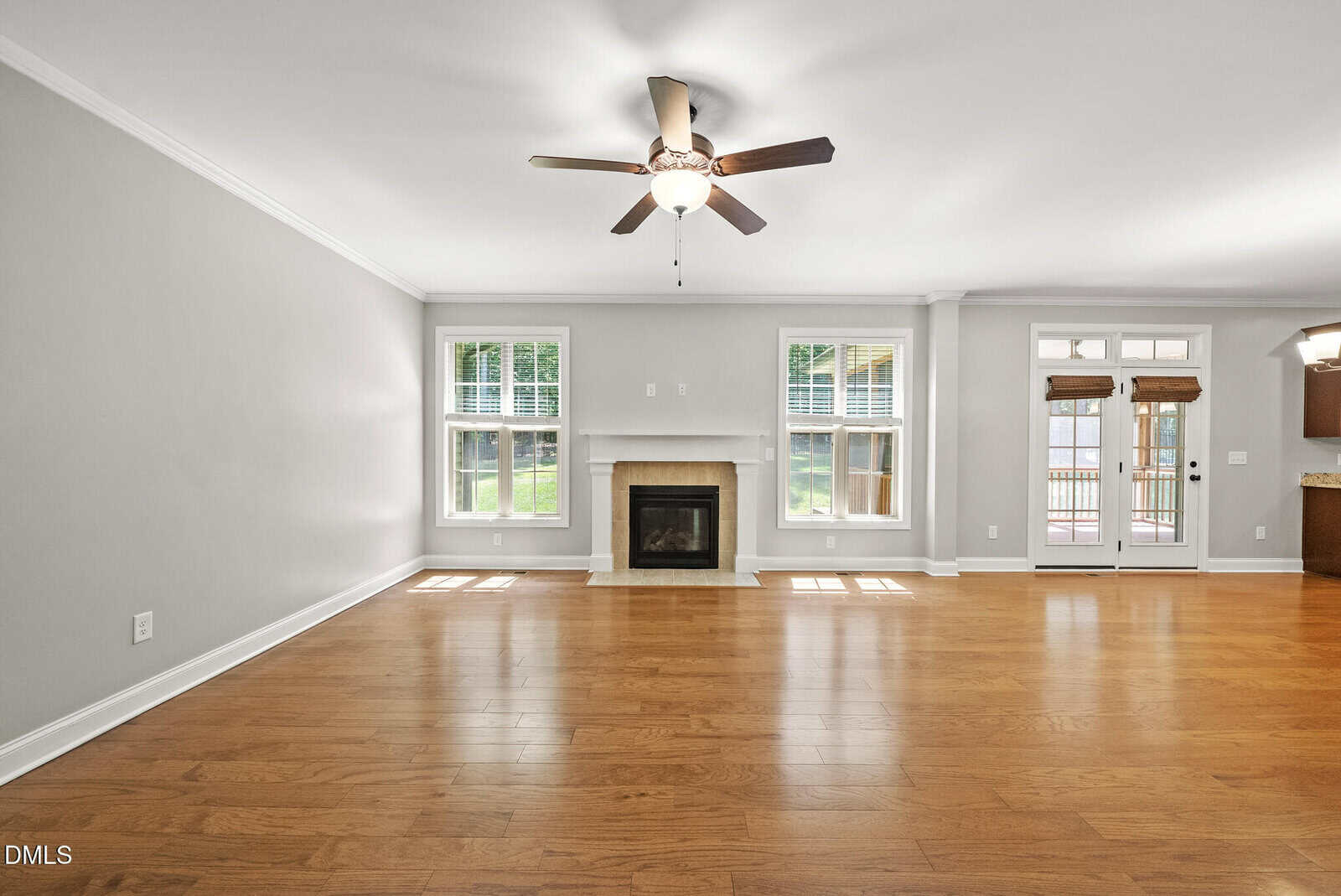 Spacious living room with hardwood floors, a ceiling fan, a white fireplace centered between two windows, and a view of a deck through French doors on the right.