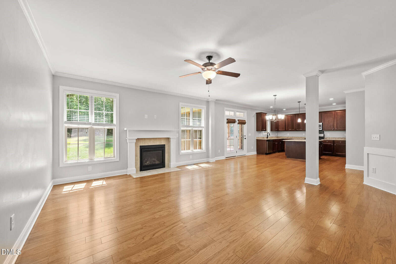 Spacious living and dining area with wood floors, ceiling fan, fireplace, large windows, and view of an open kitchen with dark cabinets and stainless steel appliances. French doors lead to a backyard.