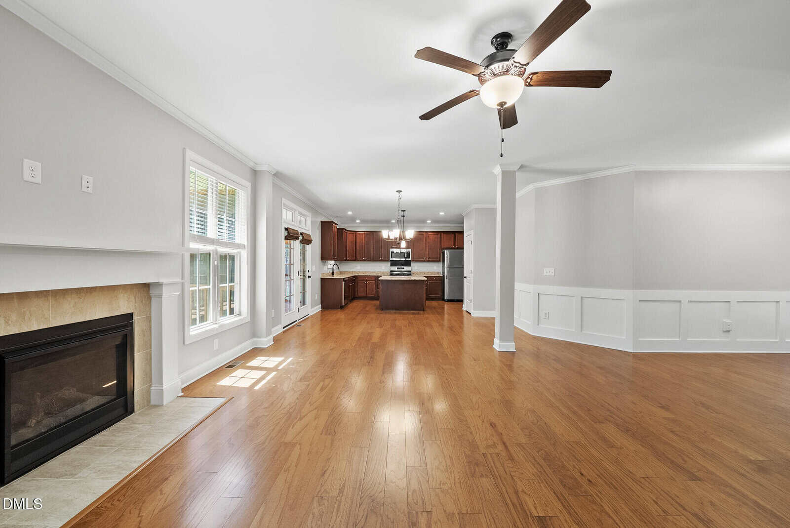 Spacious open-plan living area with wood floors, a ceiling fan, fireplace, large windows, and a kitchen with dark wood cabinets and stainless steel appliances in the background.
